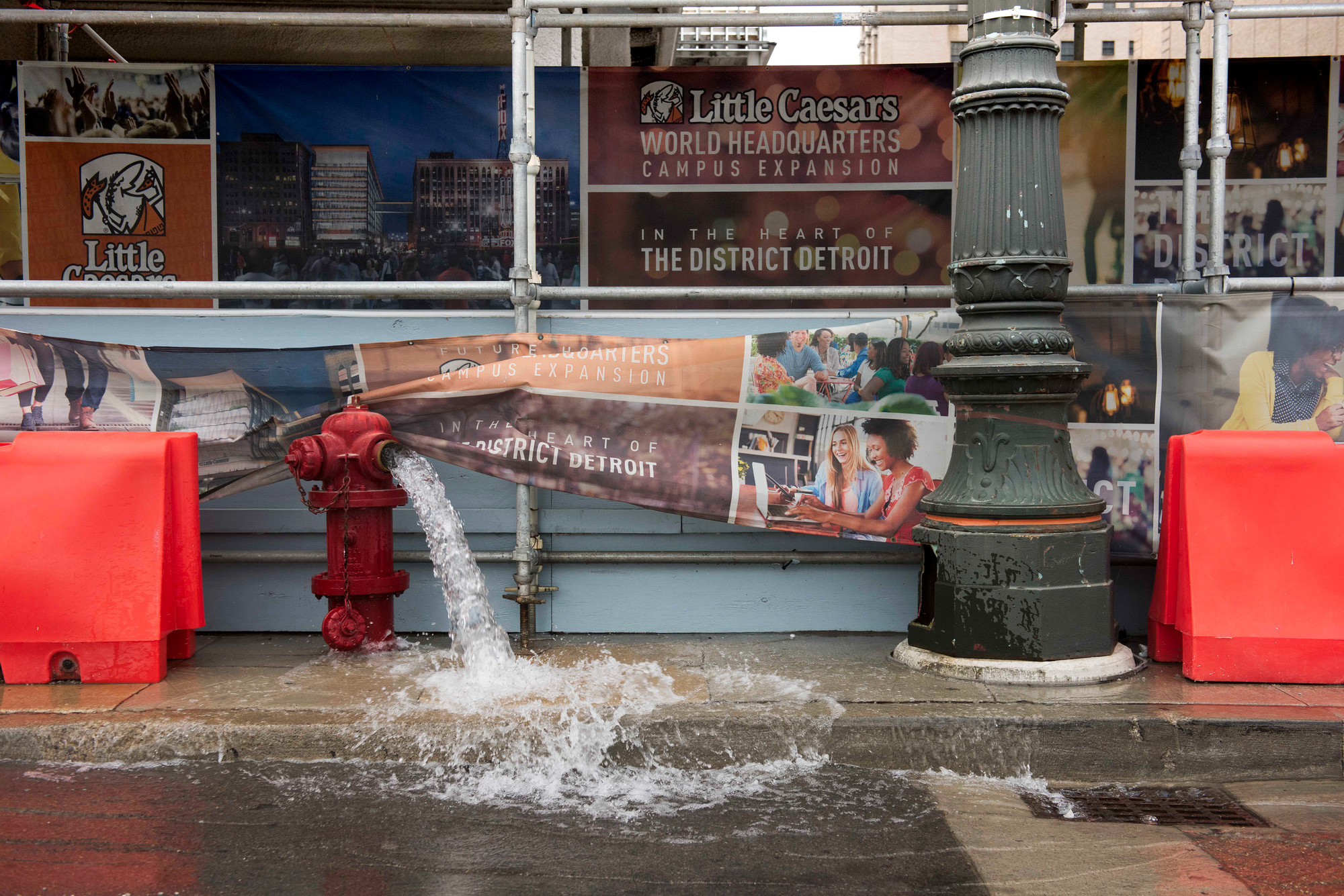 A red fire hydrant spews water onto the street in front of scaffolding and construction of the Little Caesar's Arena