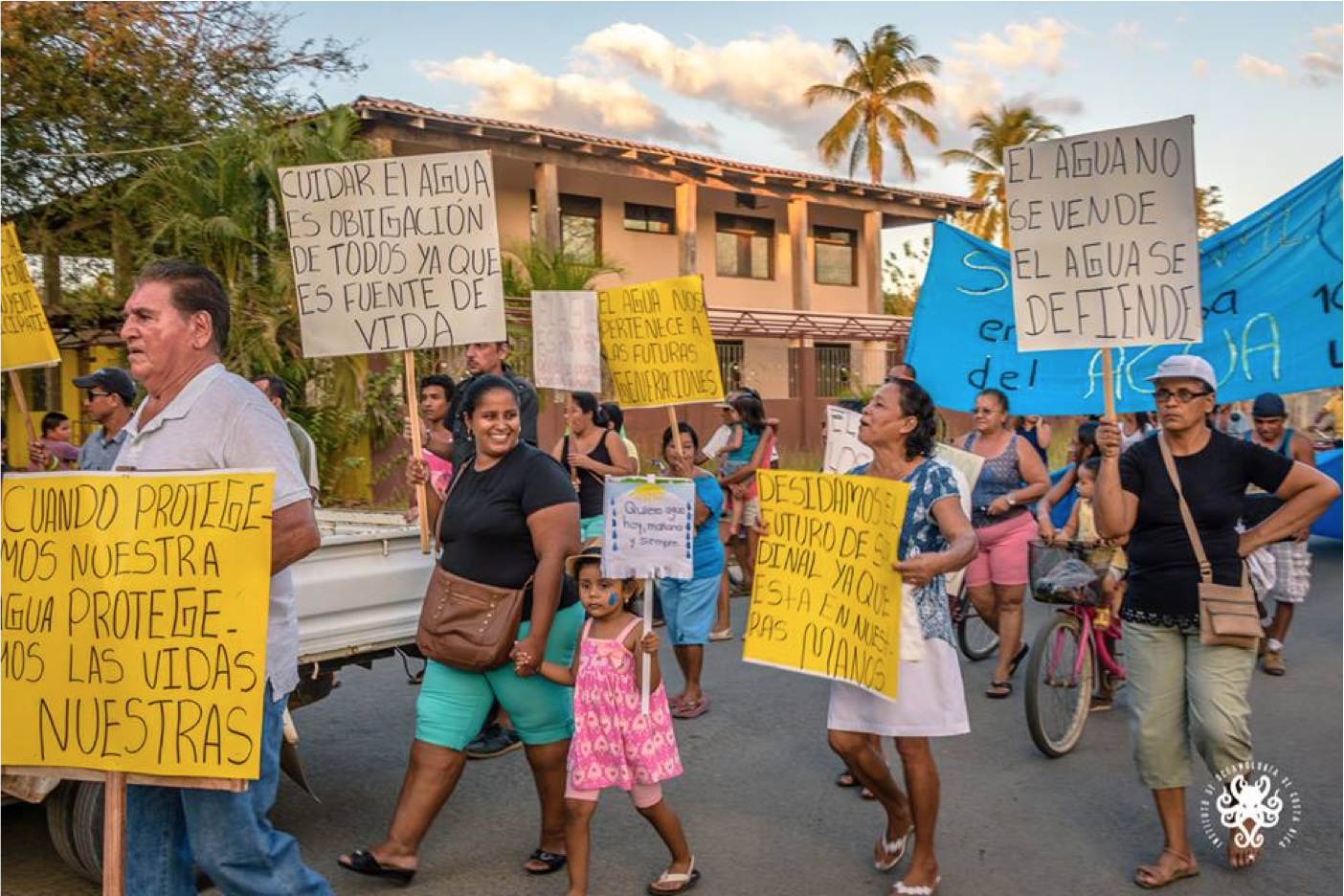 Adults and children march down a street carrying signs in a protest to protect water