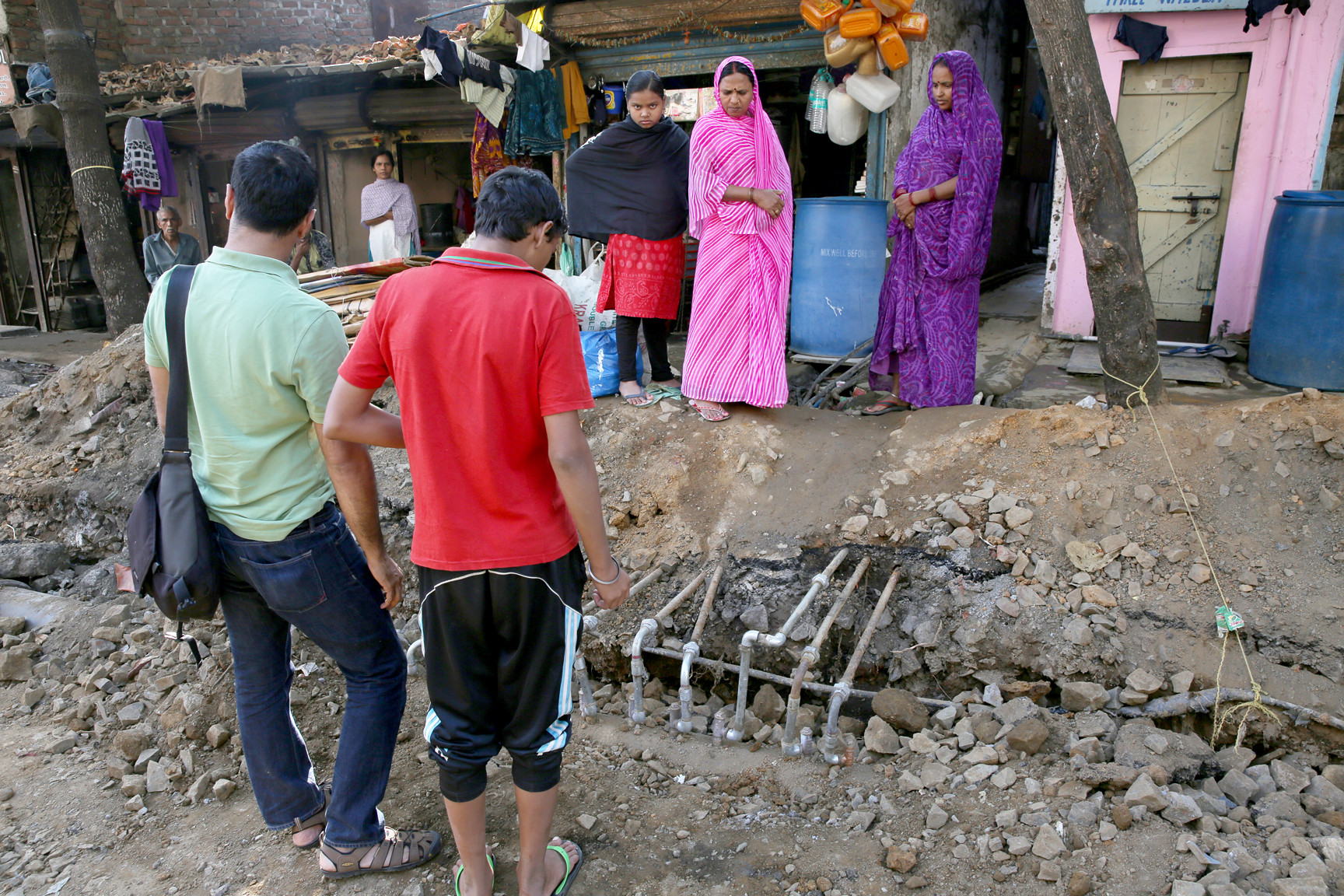 Two men and three women look at several thin pipes exposed from construction debris and dirt in front of a row of simple buildings