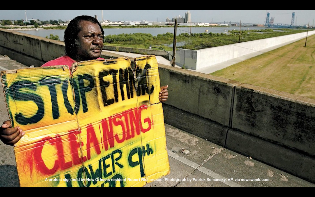 An African-American person standing on an overpass holds a hand-written sign that reads "Stop Ethnic Cleansing, Lower 9th Ward"