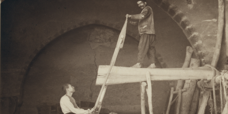 Archival photo from library of congress of two men sawing a log. One man stands on top of the log that is propped up, the other is on the ground. From Kamar ed Din Series: Hand-sawing of Logs into Planks, 1938.