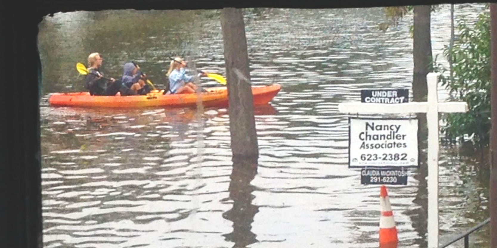 Three people on a kayak passing a for sale real estate sign in flooded area in southeastern Virginia. 