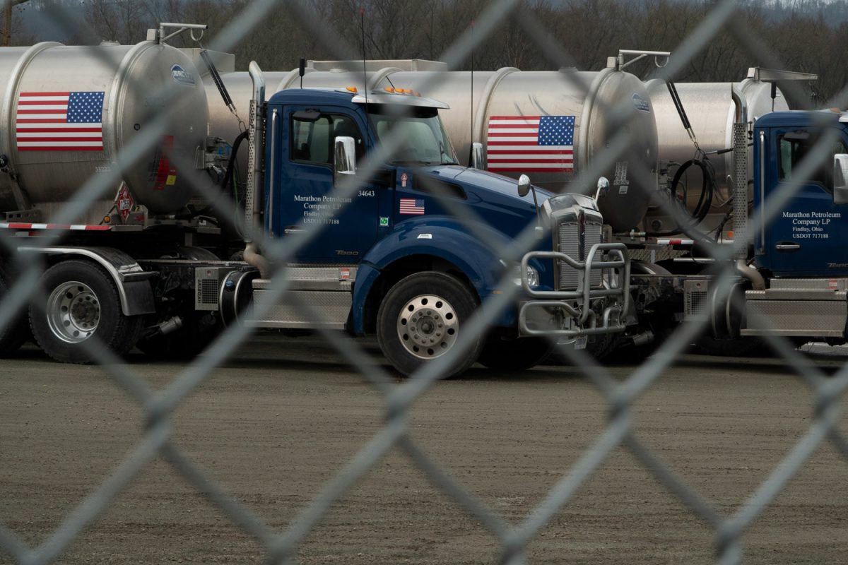 Two tanker trucks with American flags along the silver tanks and blue cabs are pictured behind a chain-link fence