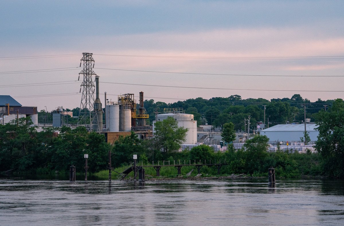 Aging industrial buildings and a large transmission tower sit behind trees on the bank of a calm river in soft morning light