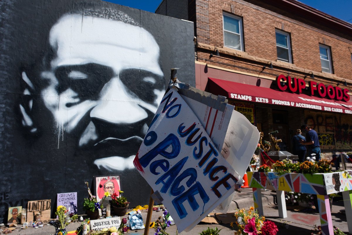 A black and white mural on the outside of a building of George Floyd with flowers, handmade memorials, and a sign reading "No Justice, No Peace" beside Cup Foods convenience store