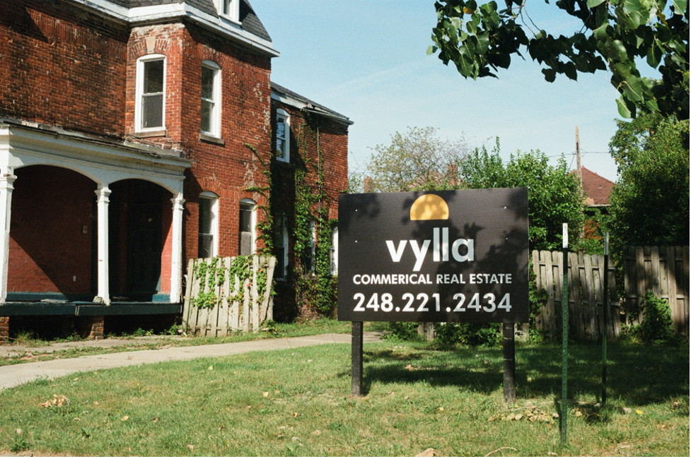 A black sign on a lawn of patchy green grass reads “VYLLA COMMERCIAL REAL ESTATE” and lists a phone number next to an older three-story red brick home with white detailing and a black gabled roof