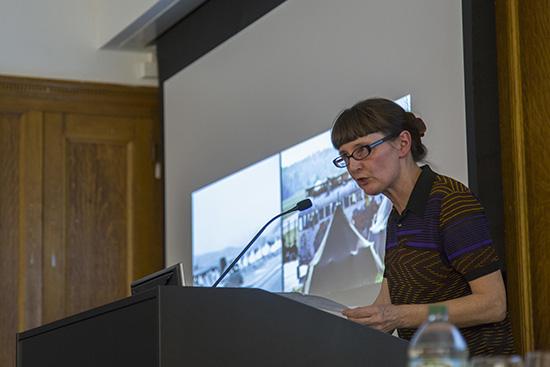 Felicity Scott speaks into a microphone at a podium in front of a projected slide showing 