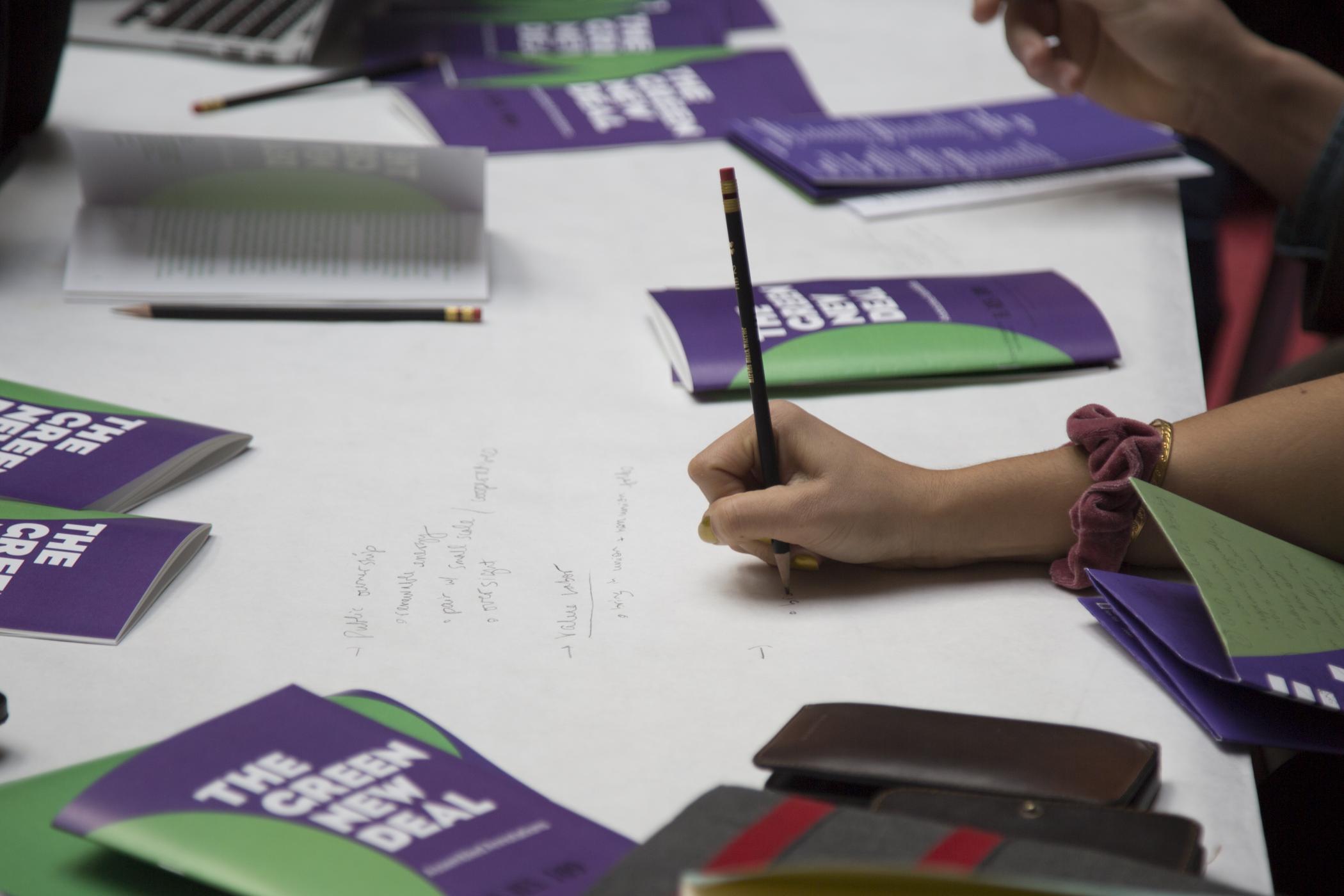 A hand grips a pencil and writes notes beside purple and green booklets titled "The Green New Deal: Assembled Annotations"