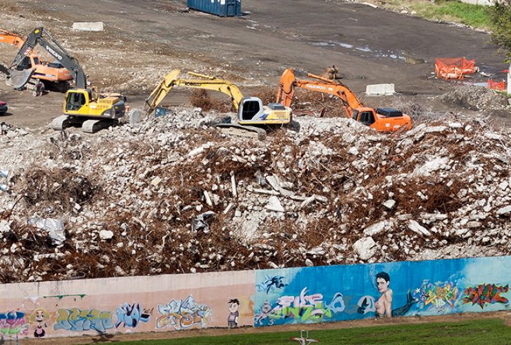Yellow and orange crane trucks sit among a pile of soil and rubble behind a wall with pink and blue murals.