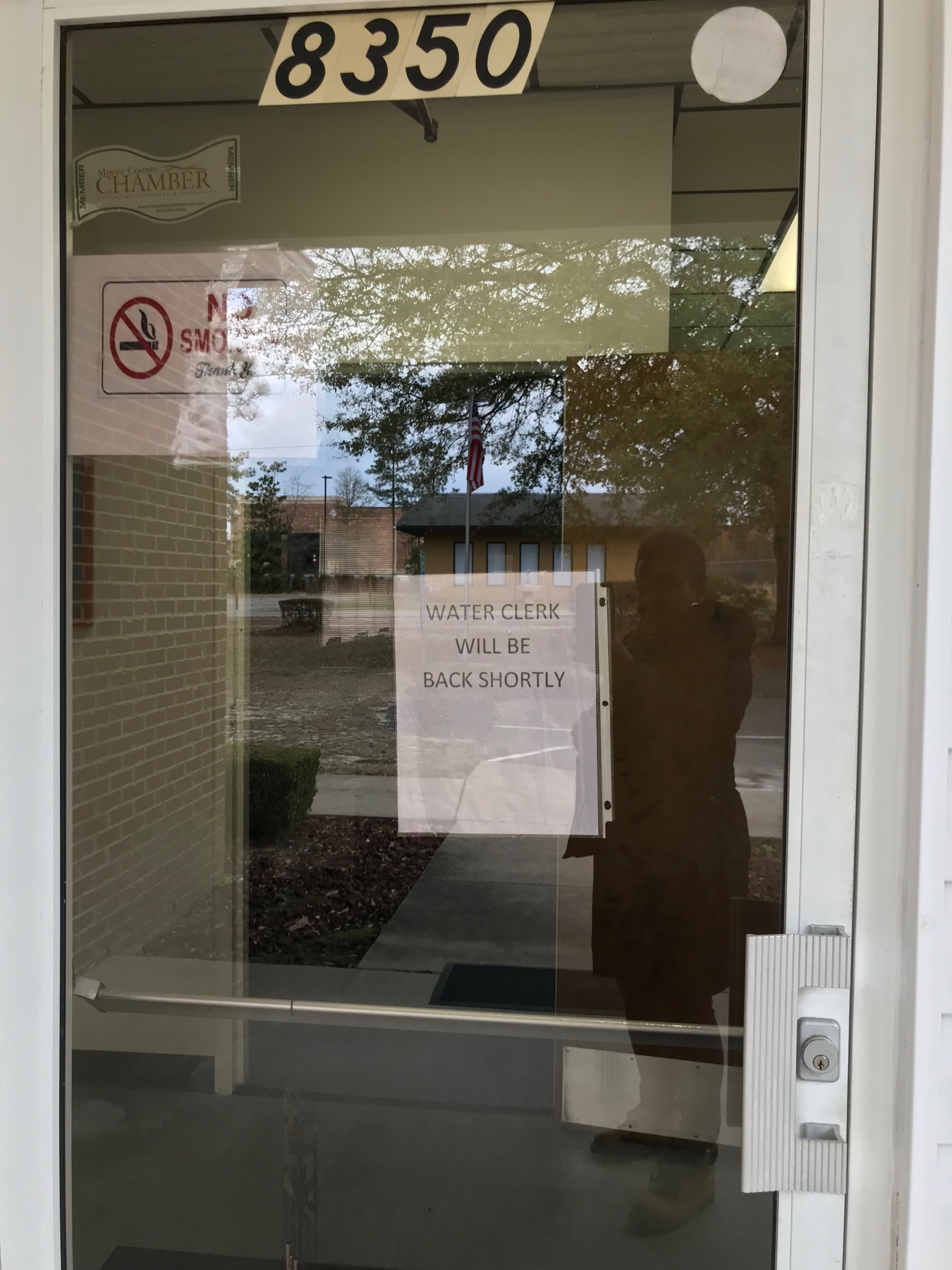 Glass door shown with typewritten sign, taped to the inside of the door, reading "Water Clerk will be back shortly." Silhouette of photographer shown in reflection.