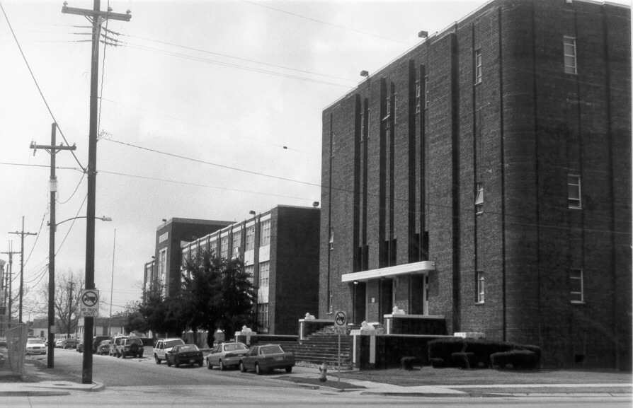 Black and white image showing cars parked alongside a two story brick structure at left, and a larger auditorium structure at right, also made of brick