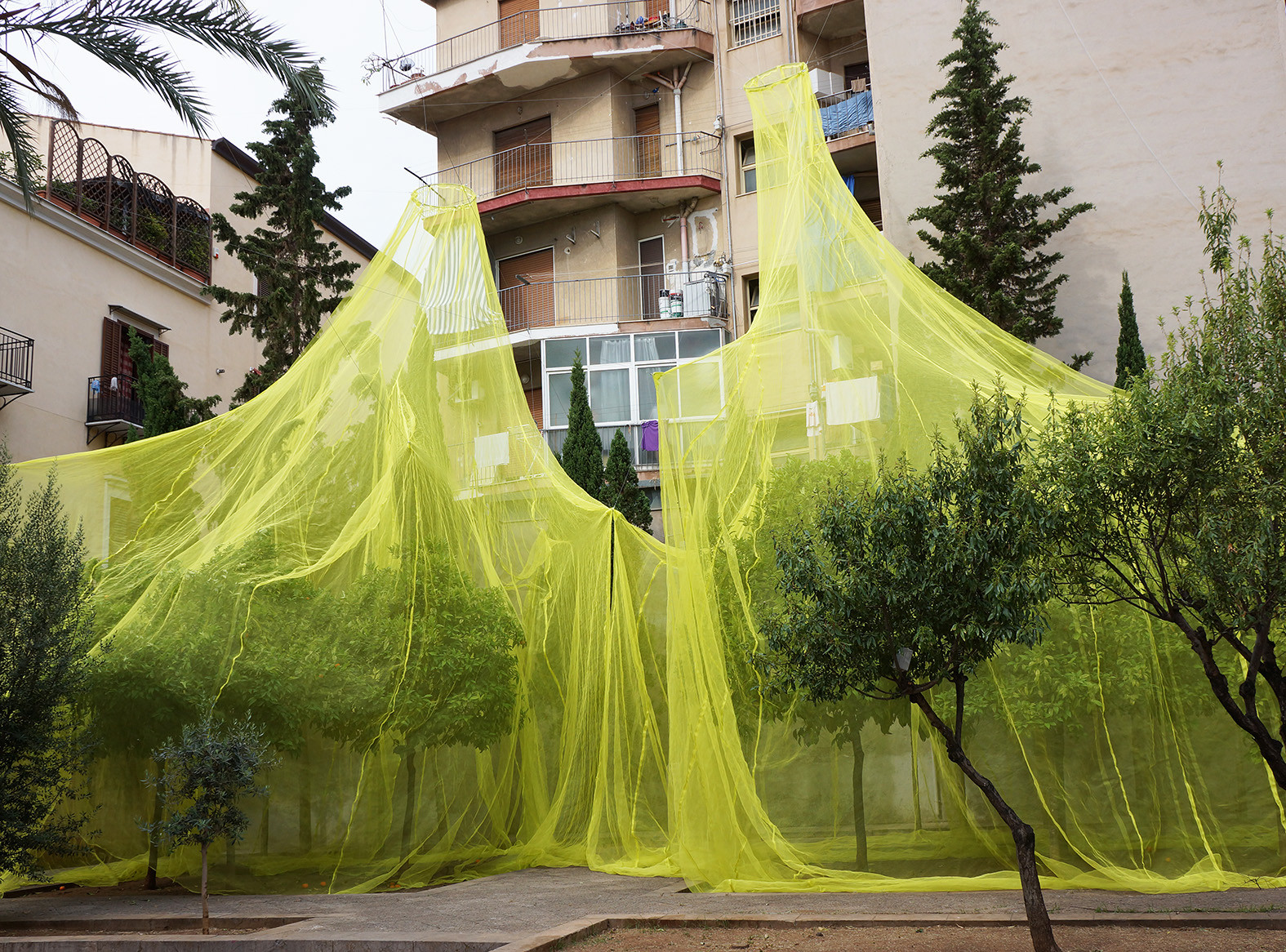 Lime green plastic netting is hung from an apartment building with several stories and balconies. The netting covers some tress while two uncovered trees stand in front of it.