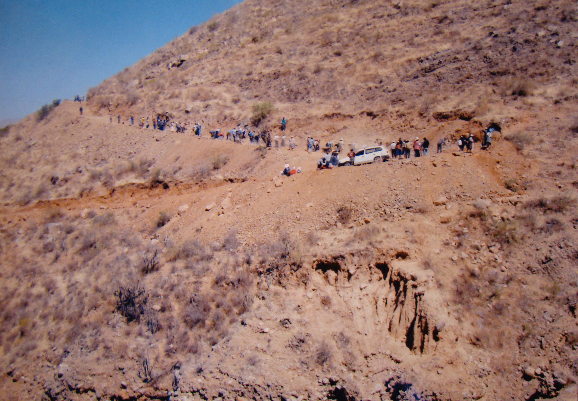 A line of people climb up a switchback on a red-brown sandy, rocky hill.