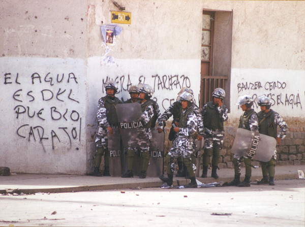 A group of armored police, wearing black-and-white camoflauge, helmets, and carrying riot shields, stand in front of a white wall with graffiti.