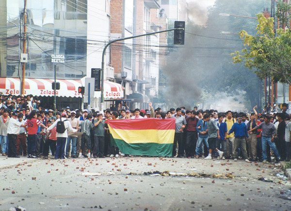 A crowd of protestors stand together, many holding hands with one another, and hold a flag with red, yellow, and green stripes. There is smoke visible in the backgroud.
