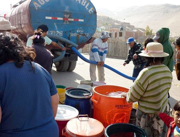 A small group of people stand among plastic tubs of various colors beside a blue tanker truck with a blue hose to fill the tubs with water.