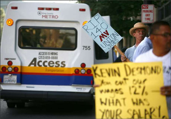 Several people stand holding homemade protest posters beside a white van with yellow, red, and blue stripes along the bottom and the word ACCESS.
