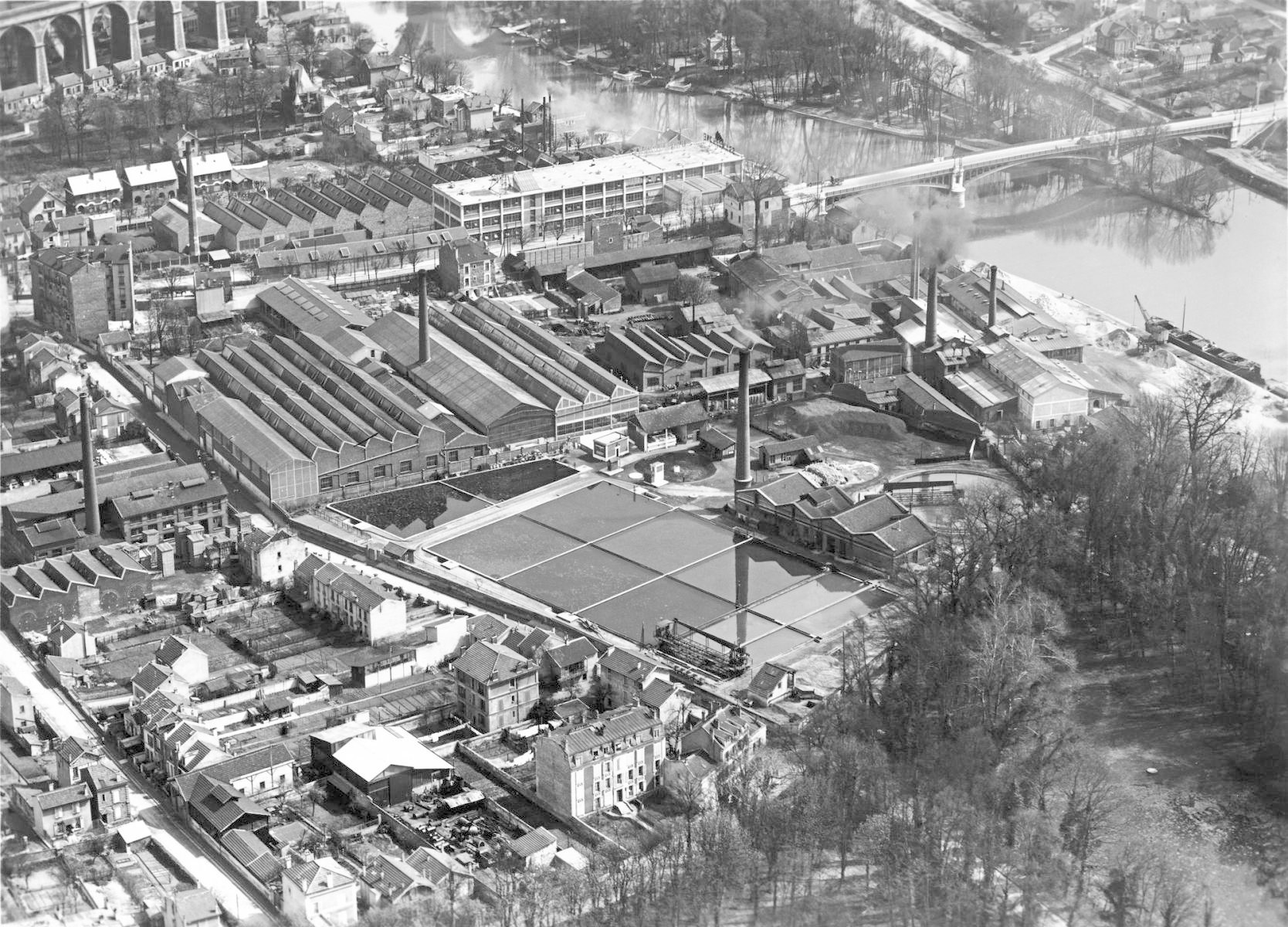 Black-and-white aerial photograph of low-slung industrial buildings with some tall, thin chimneys beside a river.