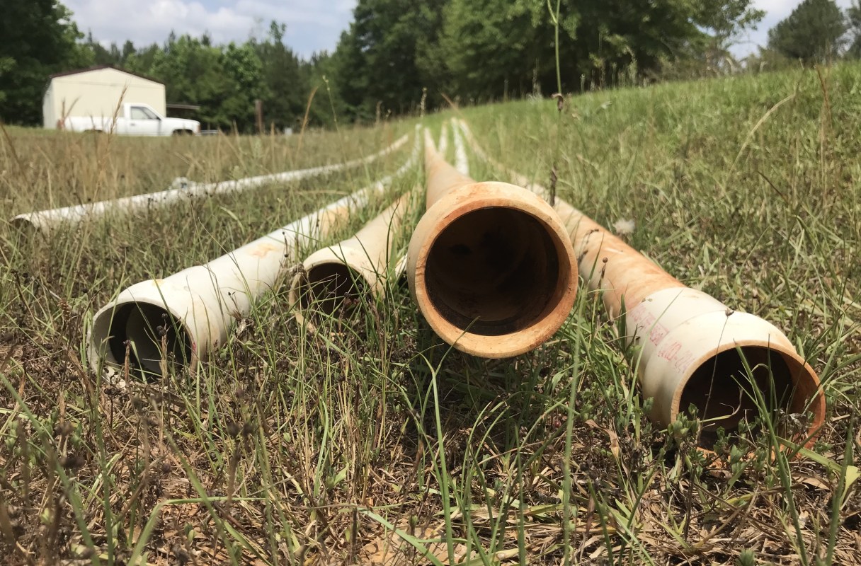 the mouths of five seemingly metal pipes are shown up close, lying in the grass, with their remainders receding into the distance. trees, a small structure, and a white truck are in the background