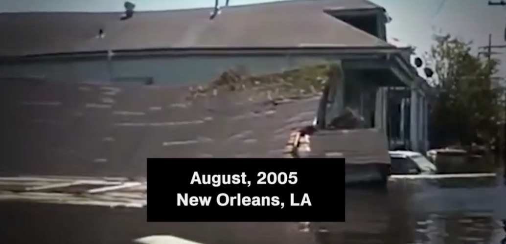 Screenshot from video showing a flooded residential street and a house with a caved in roof in New Orleans, LA in August 2005.