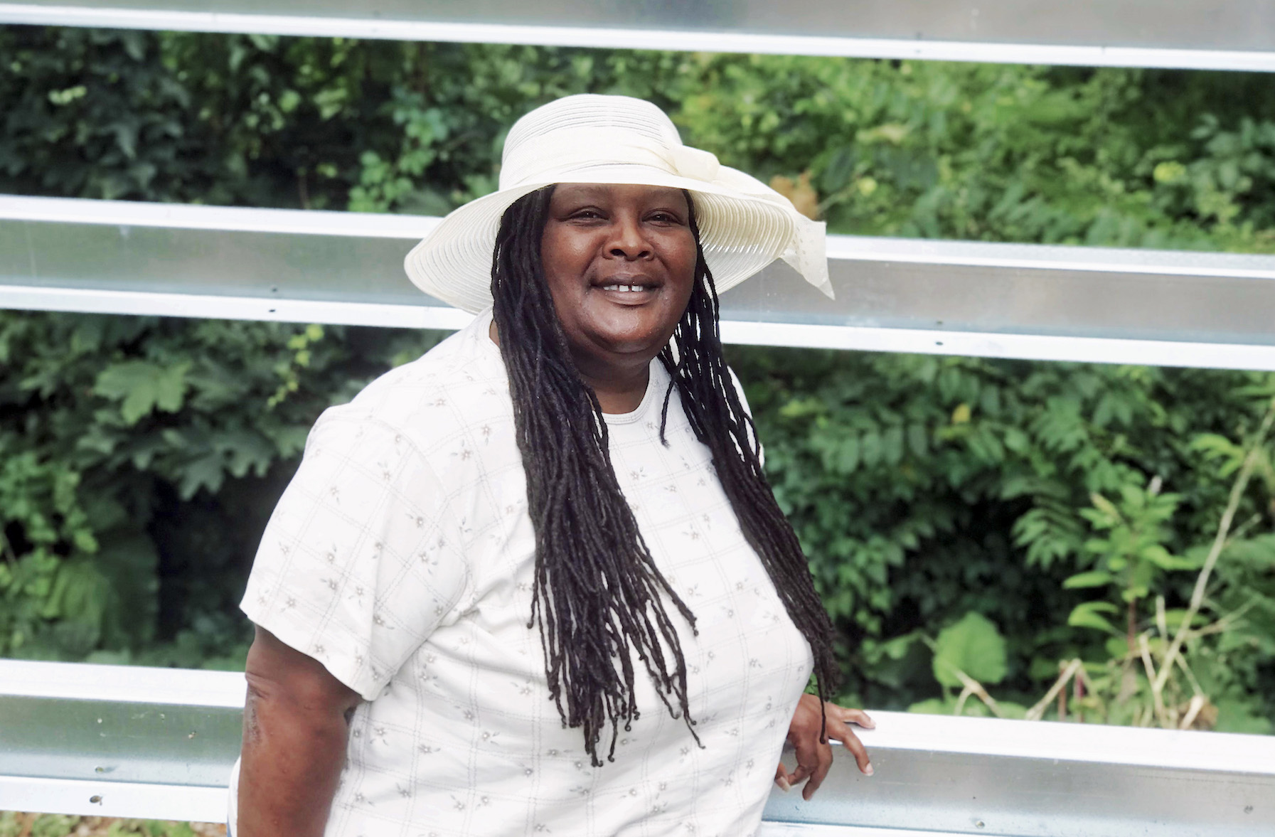 Tammi Black smiles wearing a white blouse and sunhat in front of metal bars over green bushes. Her hair is in tight braids.