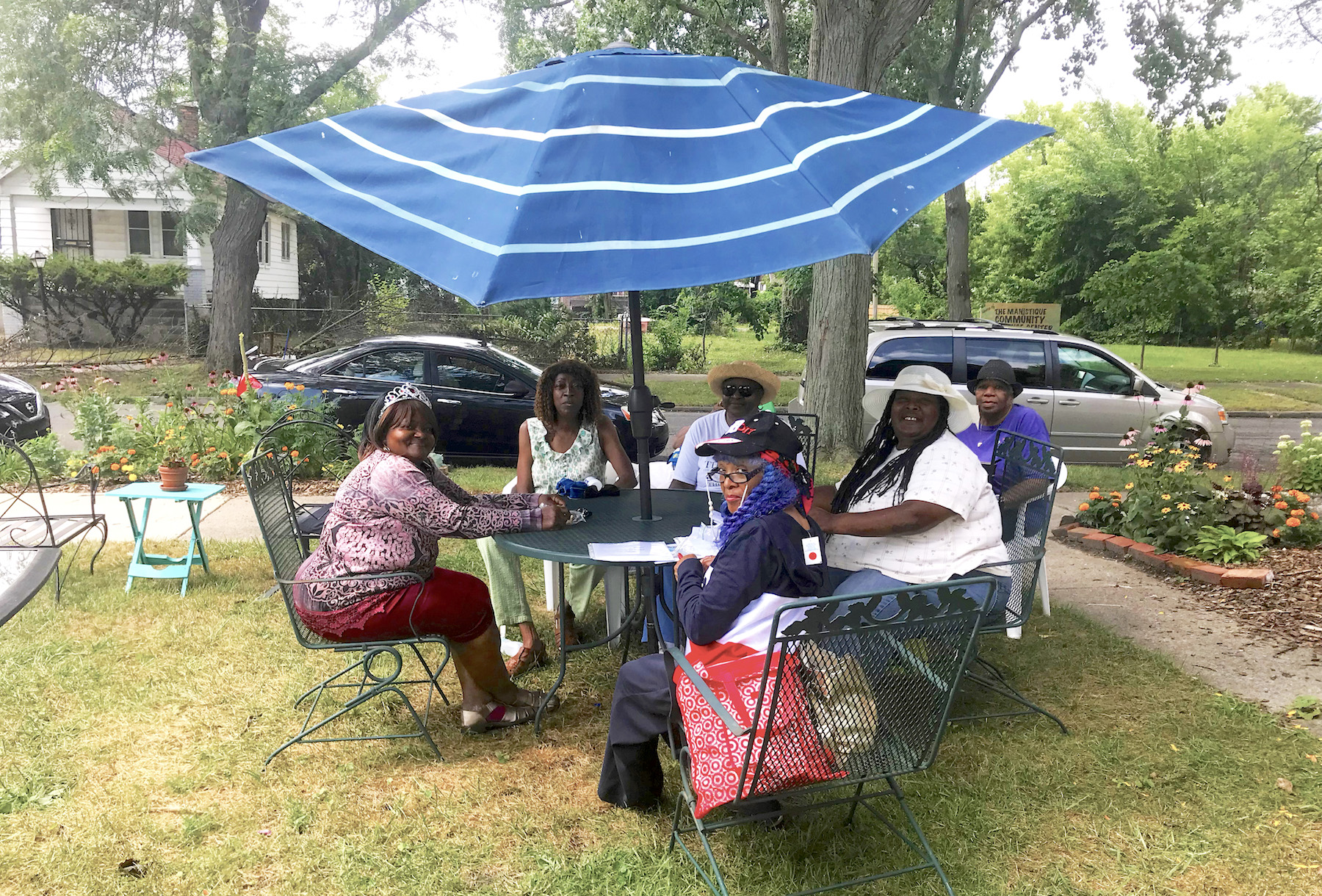 Six Black women sit around a table with big blue umbrella in a grassy yard with several trees