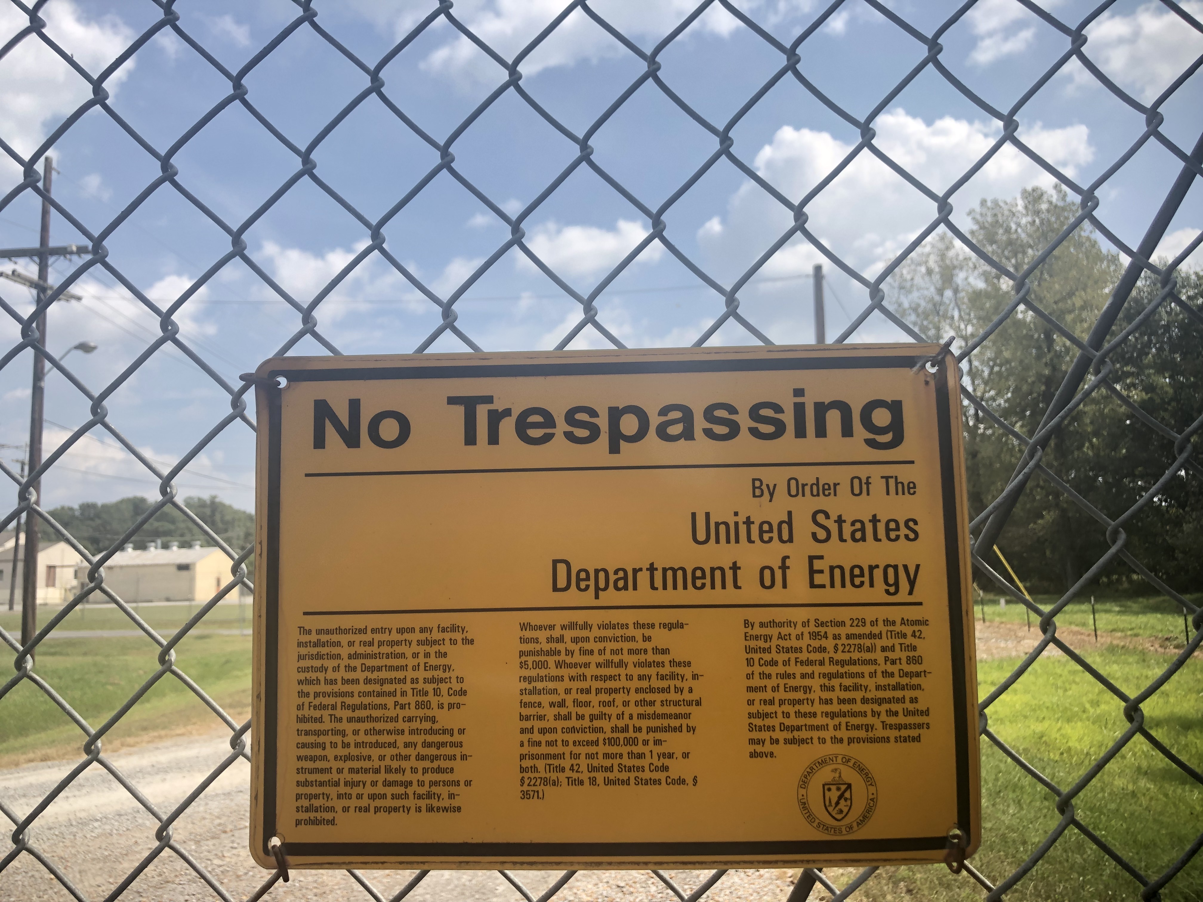 Yellow sign on fence line that reads "No Trespassing By Order Of the United States Department of Energy. The fence line separates the West Kentucky Wildlife Management Area from the Paducah Gaseous Diffusion Plant. In the distance, towers of the plant facilities are visible.