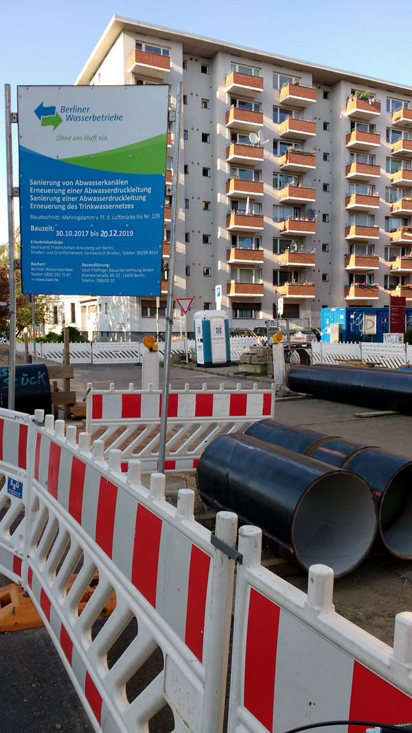 Water pipes in pieces on top of concrete waiting to be laid underground. Berliner Wasserbetriebe is in the background. Barriers with reflective surfaces surround the black pipes.