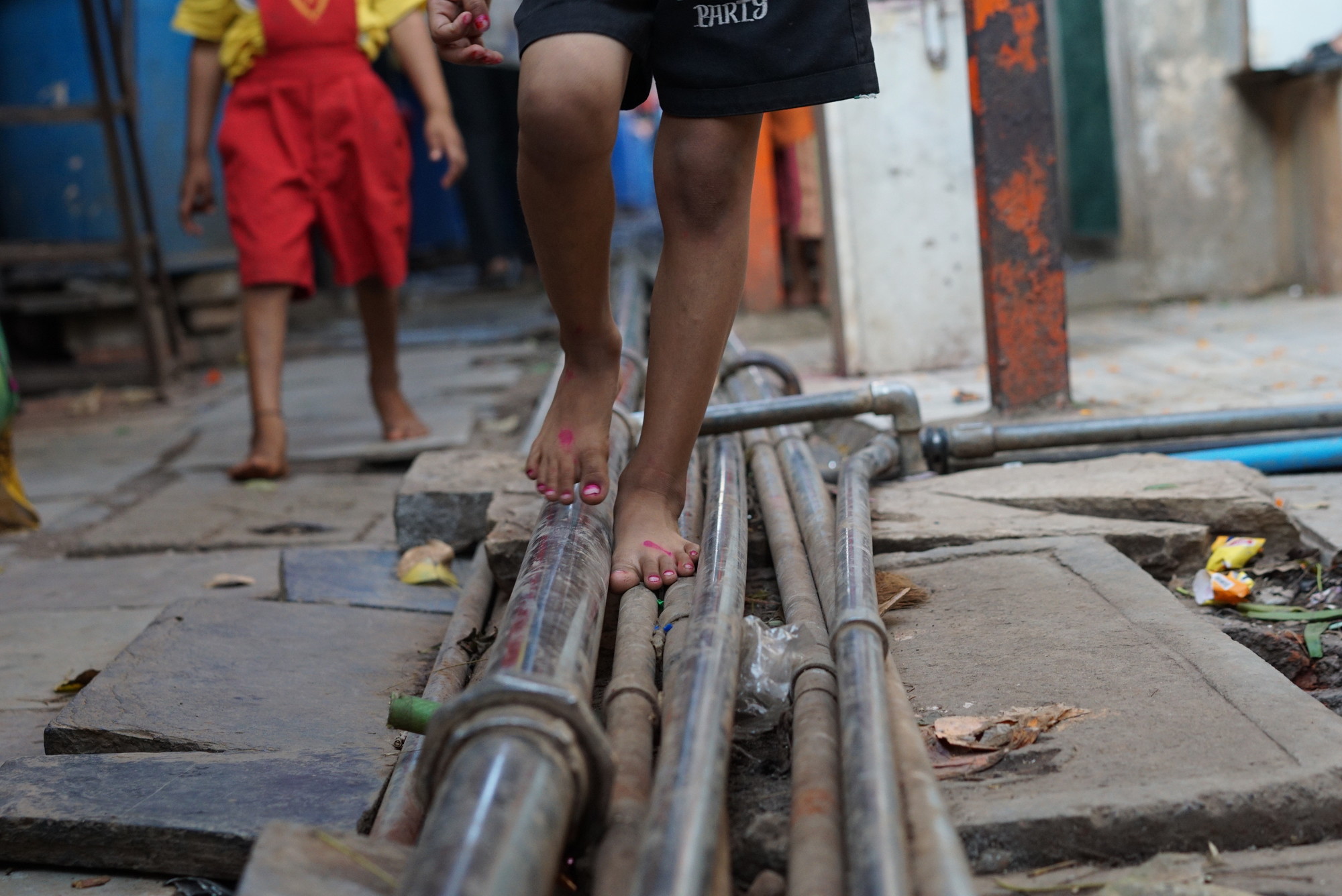 A child's thin legs, wearing black shorts, walk on top of a bundle of various-sized water pipes
