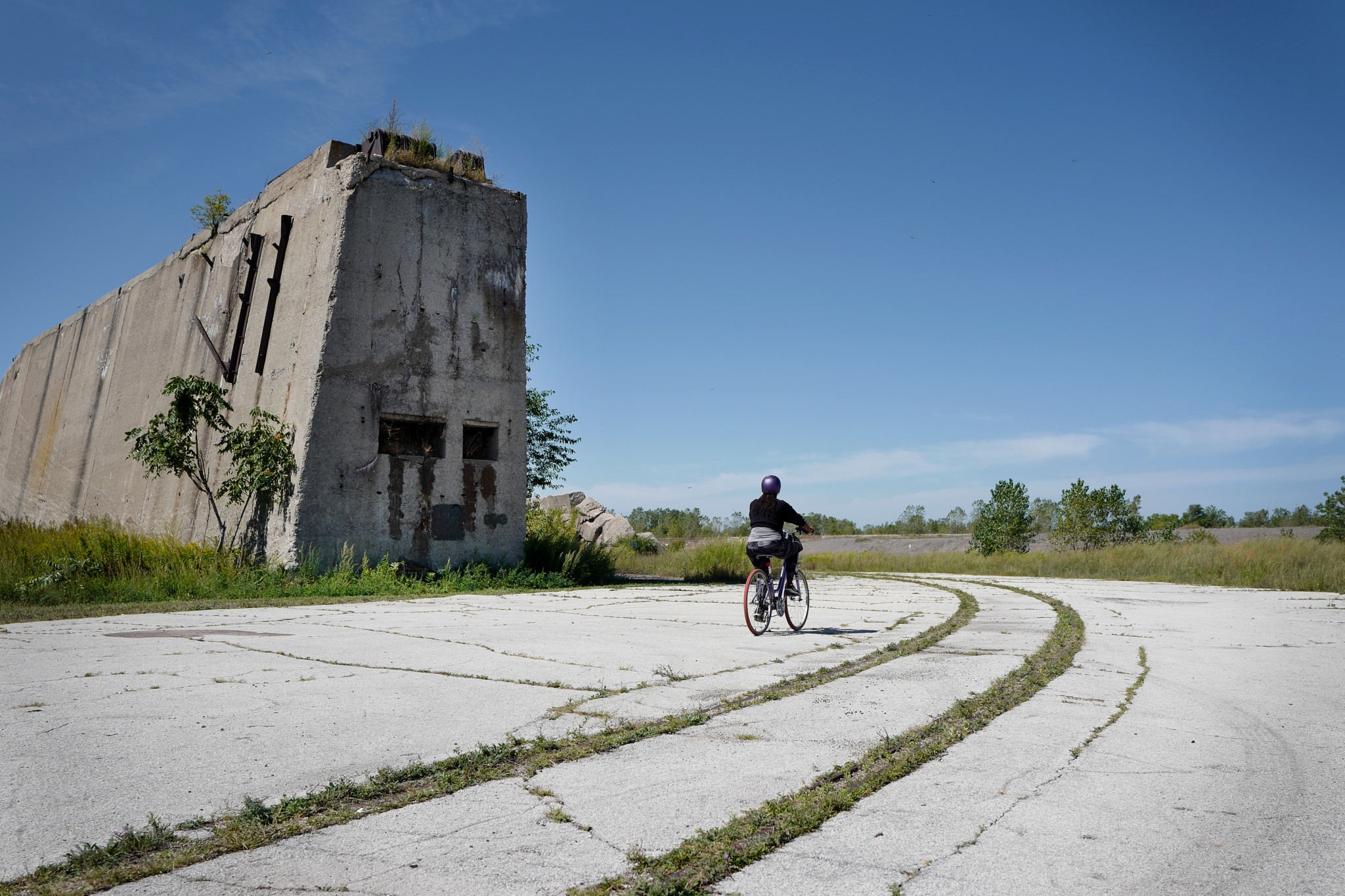 A large abandoned concrete structure sits beside cracked concrete paths on which a bicyclist rides towards a blue horizon, with tufts of grass and shrubs