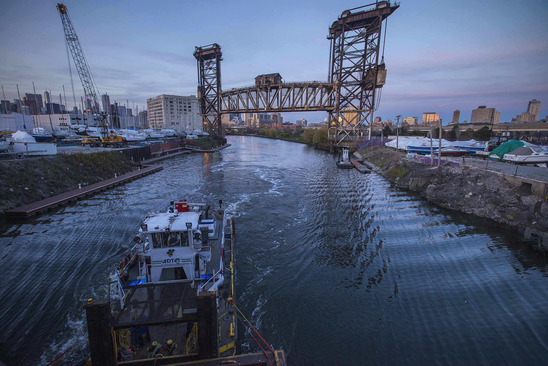 A boat moves along a dark river through an industrial landscape with a steel vertical lift bridge in the background