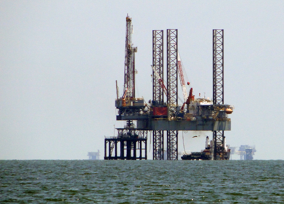 An oil rig rises out of the blue-green water of the Gulf of Mexico with other rigs or ships in the distance. The photo is slightly out of focus as if from heat distortion
