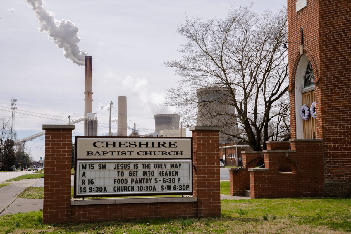 The Cheshire Baptist Church sign reads "Jesus is the only way to get into heaven" and advertises worship times and a food pantry next to a simple red brick church. In the background smokestacks spew white clouds into the gray blue sky