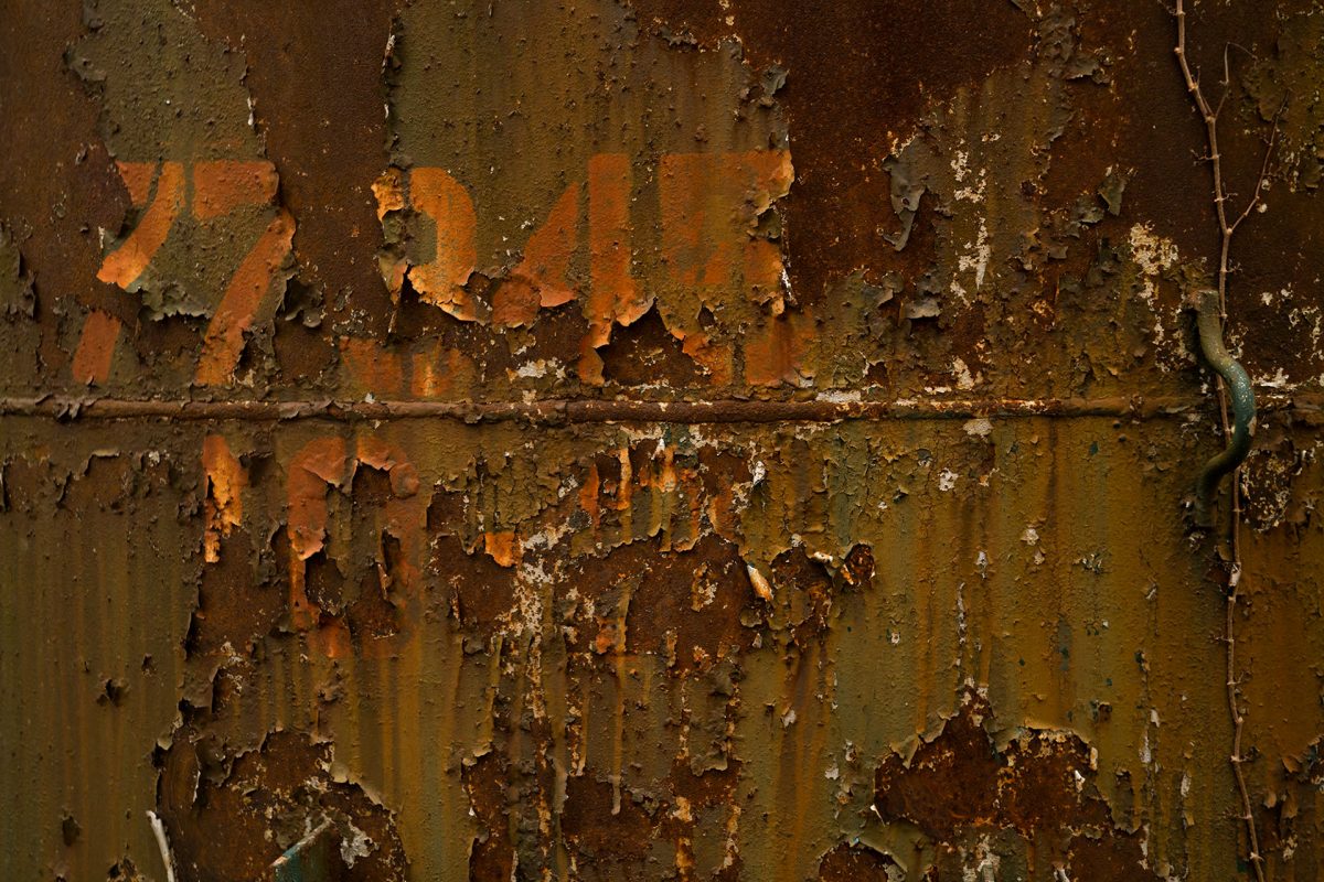 Layers if olive paint peel off of an unidentified rusting metal surface that could be an oil barrel