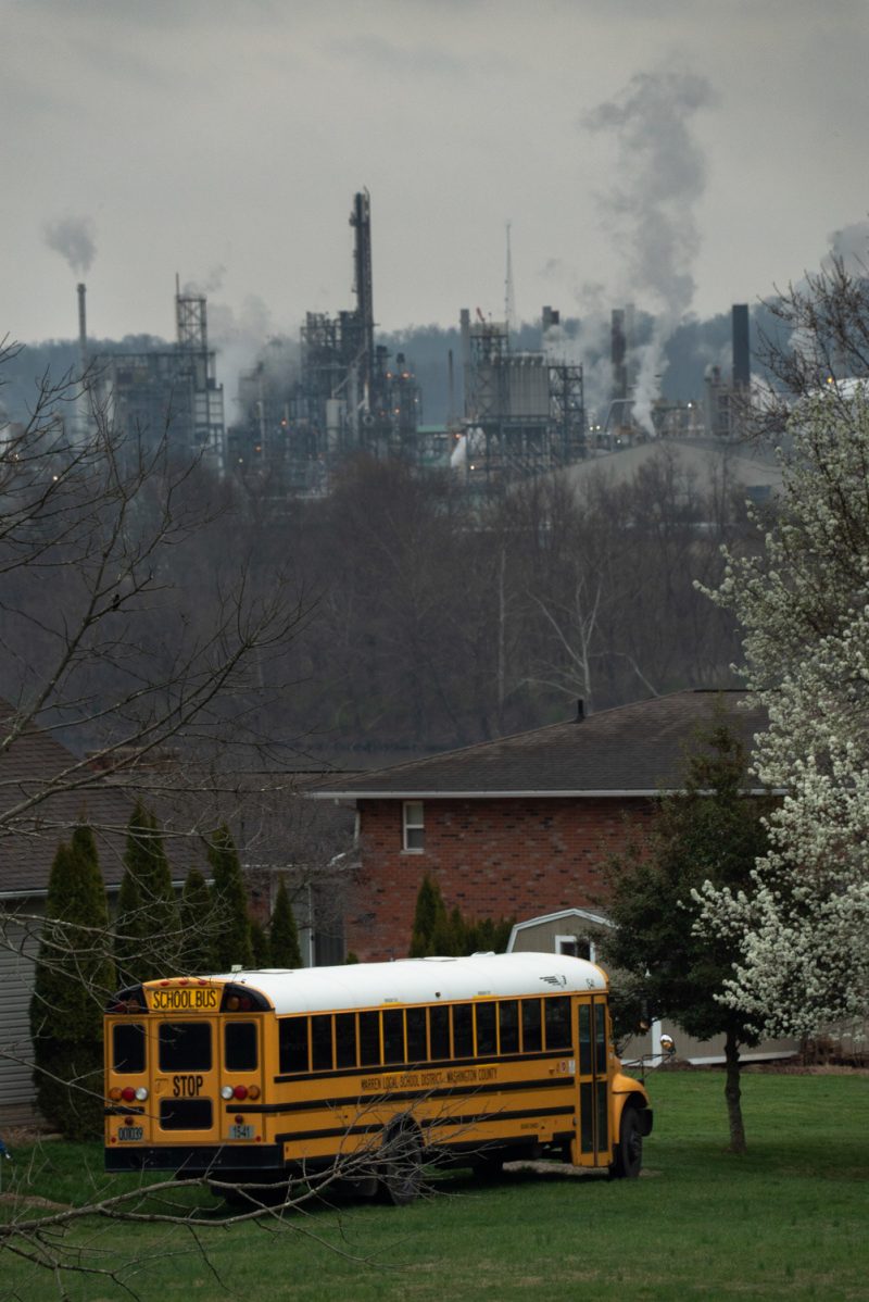 A yellow schoolbus is parked in a grass yard in front of a red brick house and leafless trees with an industrial plant spewing pollution in the background