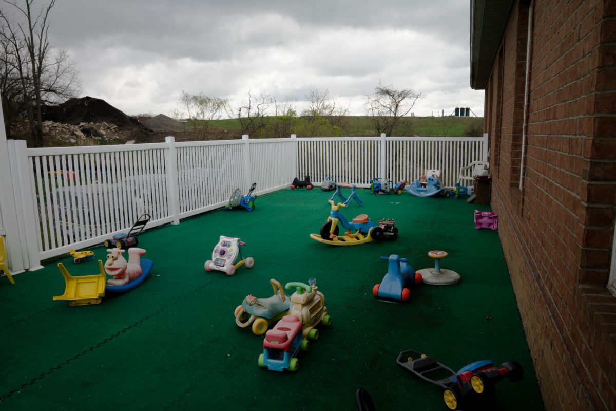 Children's toys are strewn about a green turf area next to a brick house with a white fence. The sky is cloudy.