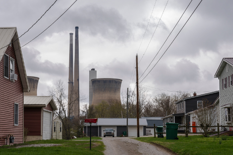 A cooling tower and smokestacks are seen in the background with a dirt road lined by modest two-story homes in the foreground. The sky is cloudy