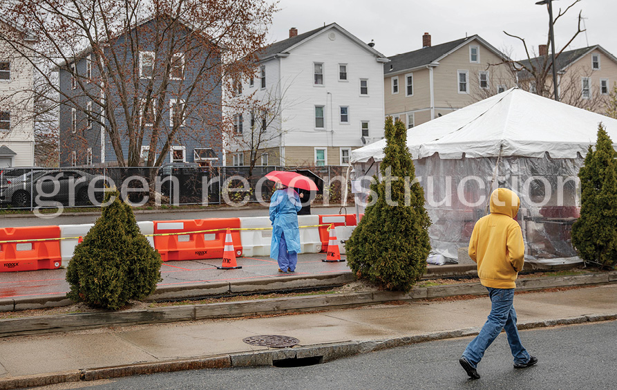 Orange barricades and covered faces punctuate an outdoor scene with grey residential architecture in the background and temporary shelter in the foreground.