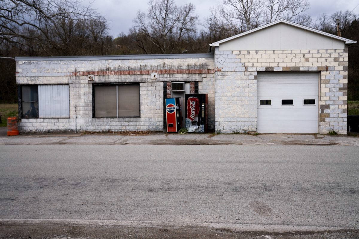 An abandoned single-story white building with a white garage and two old vending machines photographed from across the road