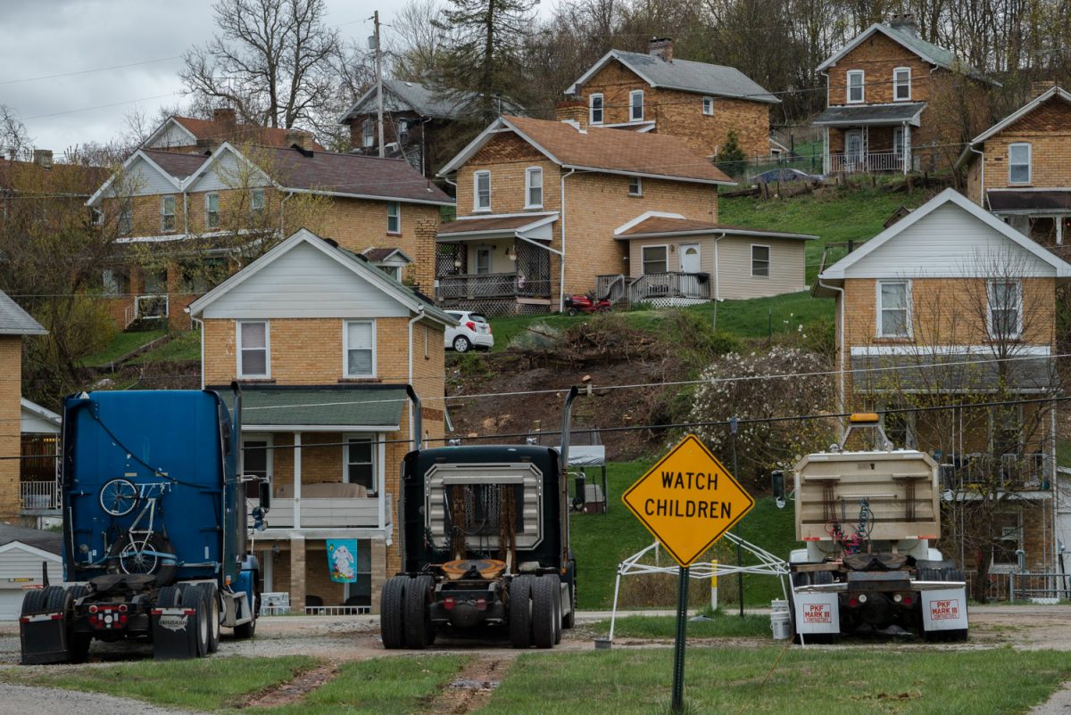 A number of simple, two-story homes with pitched roofs are seen along a hillside with three trailerless truck cabs and a sign saying "WATCH CHILDREN" in the foreground