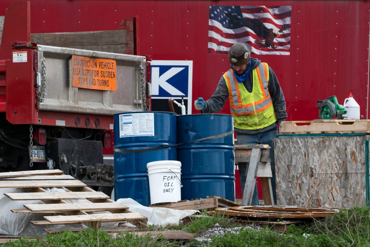 A man wearing a hardhat and reflective vest works beside two blue metal barrels in front of a red wall with an American flag decal