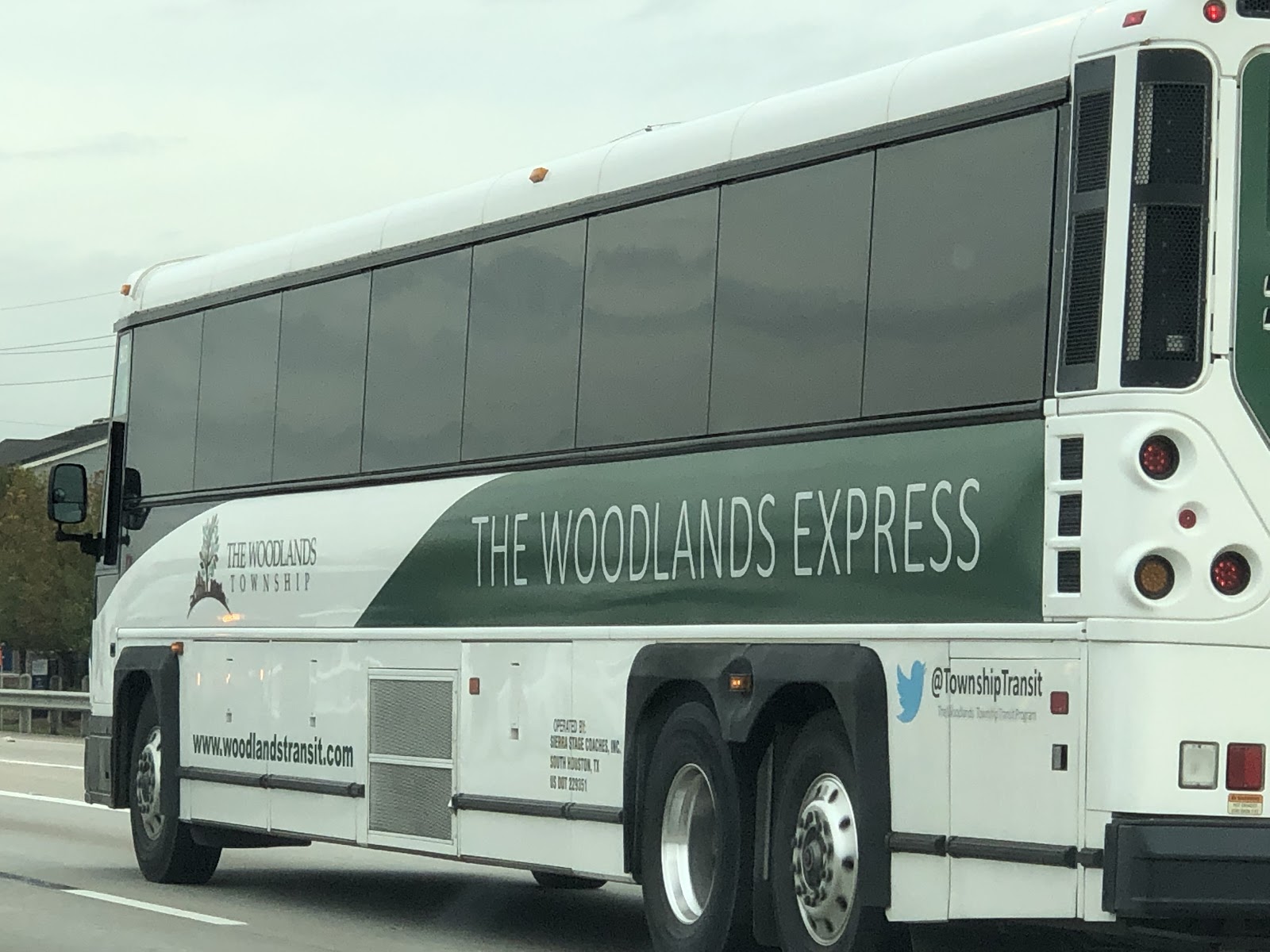 A white passenger bus with tinted windows and a green bar beneath with white text overtop reading "The Woodlands Express"