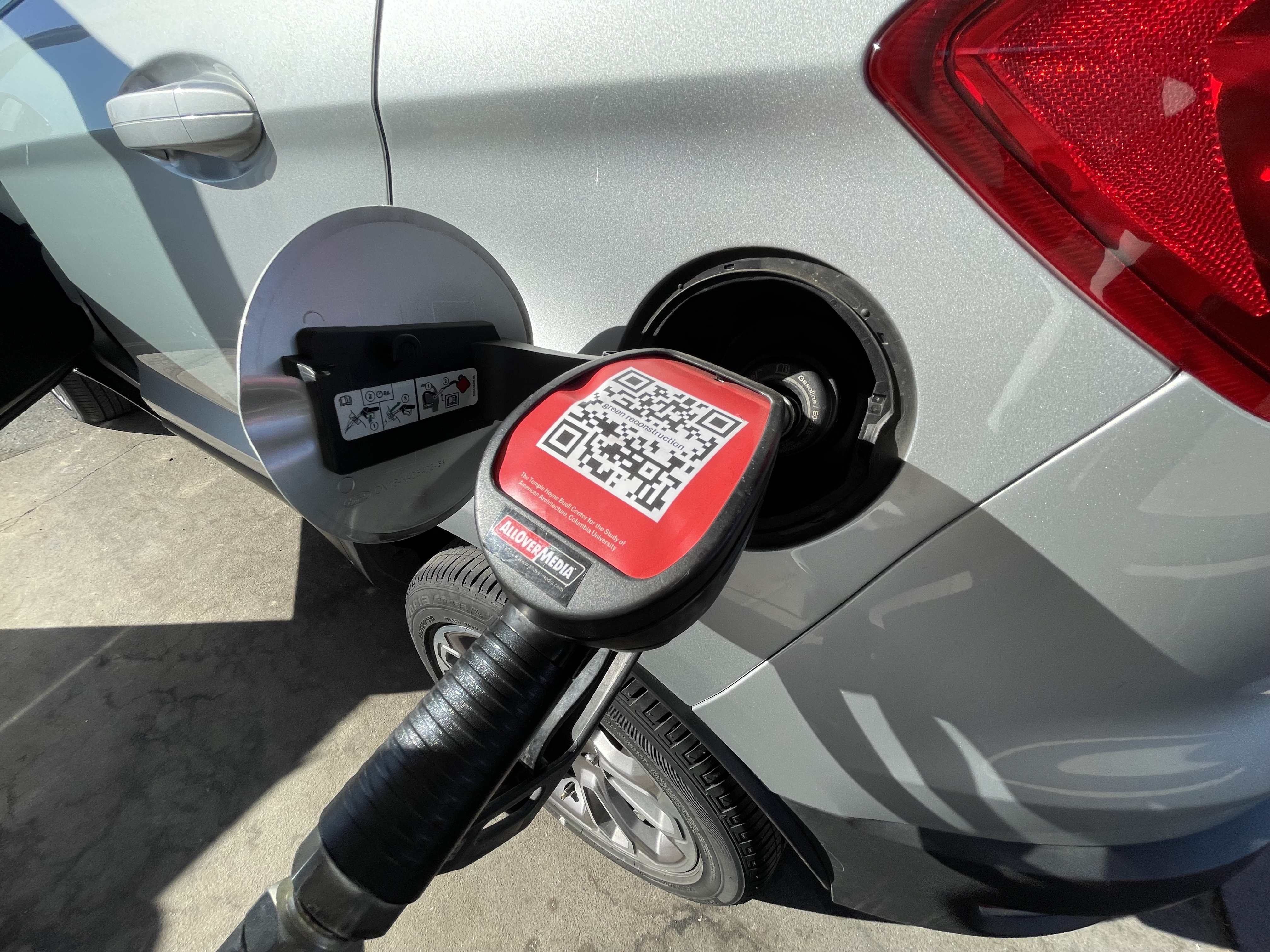 A gas pump pumping gas in a silver car. On the handle a A bright red frame surrounds a QR code