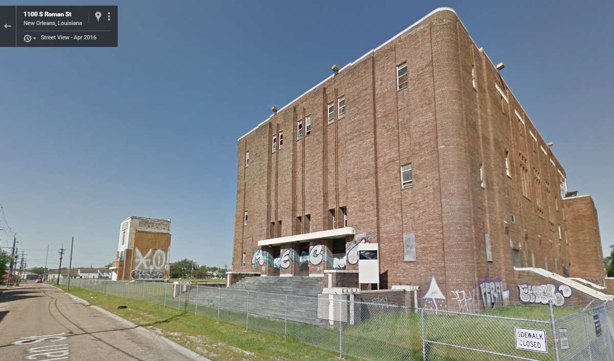 A Google Streetview image shows a brick auditorium, right, behind a chain link fence, alongside an open field and a small brick structure in the distance.
