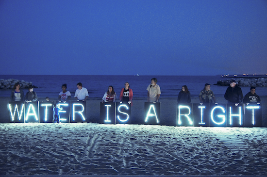 People stand in a line on the sand in front of open water at night holding white neon letters spelling "WATER IS A RIGHT"