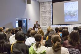 Photo from the back of the audience showing people looking on as Kim Stanley Robinson stands behind a podium to speak with his book cover projected on a screen beside him