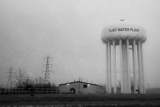 A water tower with "Flint Water Plant" painted on it is shown against a grey sky in a hazy black-and-white photo