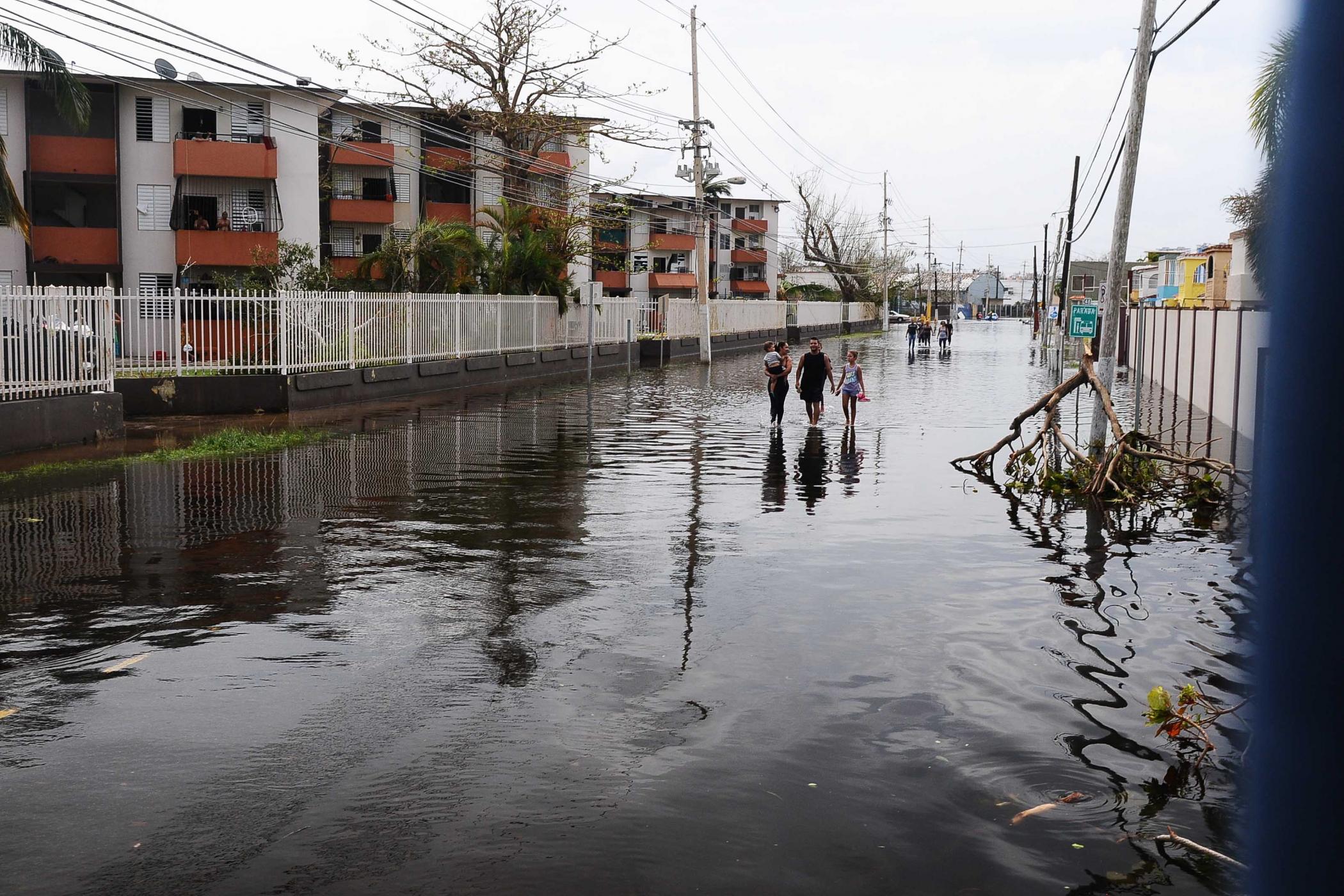 Three people walk hand-in-hand through knee-deep water on a flooded street with power lines overhead on an overcast day.