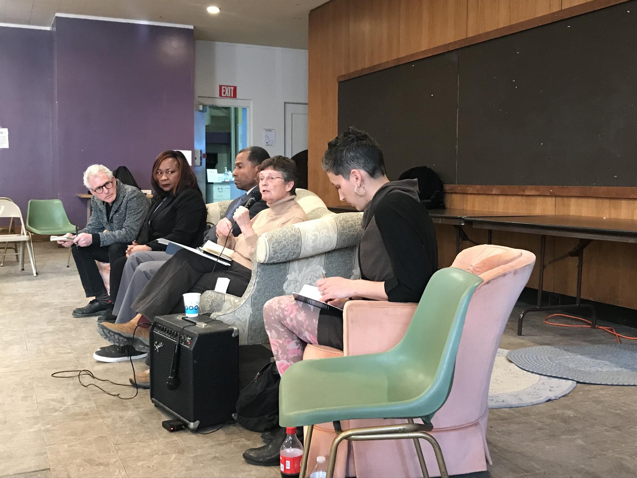 Five discussants, on in a pink armchair and four on a gray and white couch, sit in a line in front of a blank chalk board. One participant holds a microphone to speak while the others look at her or their own notes