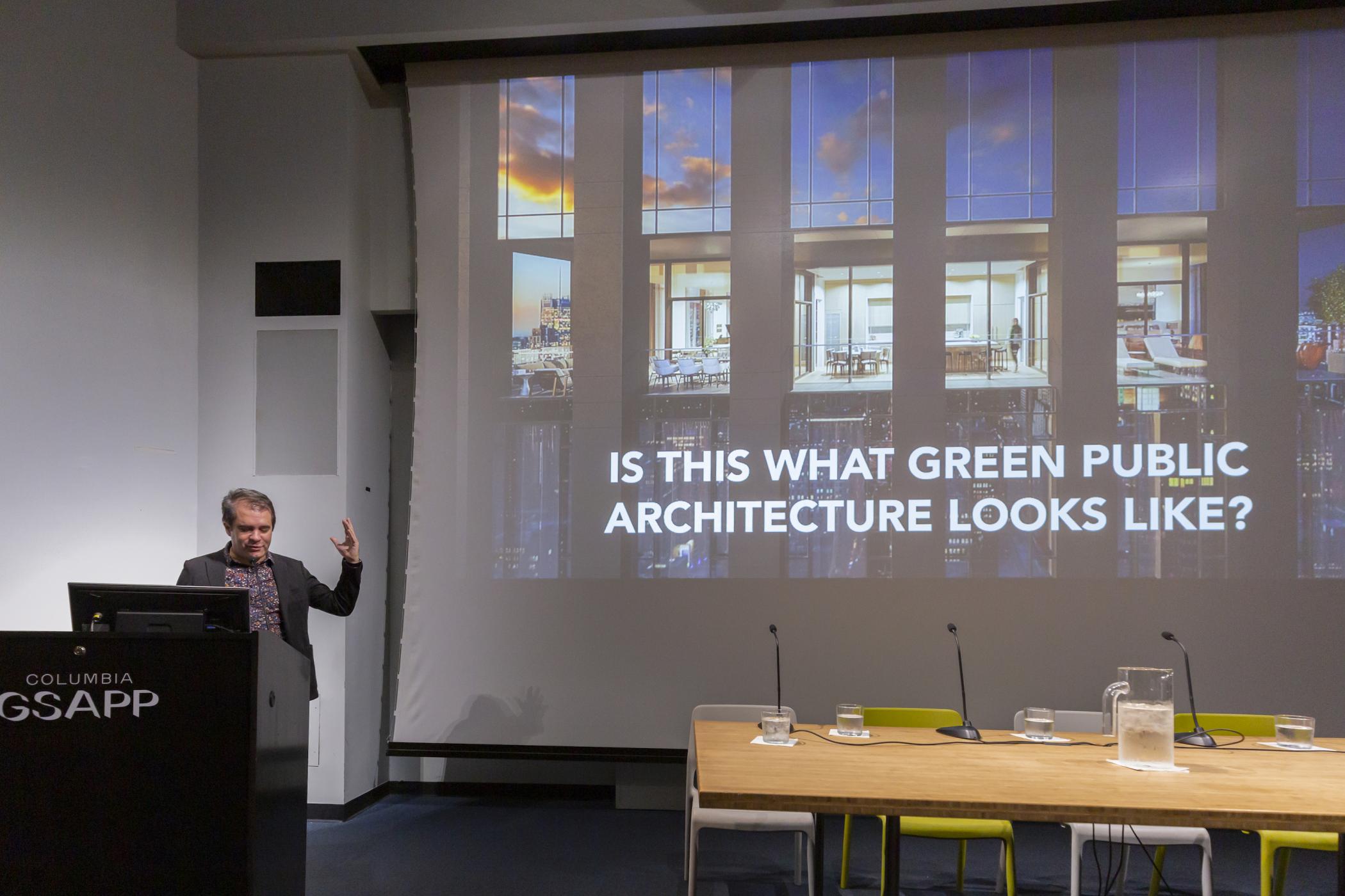 Andres Jaque stands at the podium gesticulating in front of a slide of a modern glass building with text saying "is this what green public infrastructure looks like?"